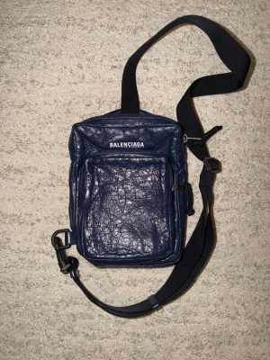 Mörkblå axelväska från Balenciaga - Säljer en Balenciaga axelväska/crossbody i mörkblå färg. Väskan är knappt använd och i mycket fint skick, med klassisk Balenciaga-logga framtill, justerbar axelrem, dragkedjestängt huvudfack samt ytterficka framtill. Perfekt storlek för det viktigaste och smidig att bära till vardags. Insidan är ren och väskan kommer även med original dustbag. Made in Italy. Serienummer syns på innerlappen enligt bild.