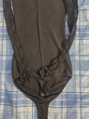 Svart body med spetsärmar - Snygg svart body med långa spetsärmar och tryckknappar i grenen. Kroppsdelen är i mjukt tyg medan ärmarna är i transparent spets med blommönster. Perfekt för att skapa en stilren och trendig look.