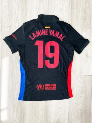FC Barcelona Lamine Yamal matchtröja - Snygg svart FC Barcelona matchtröja från Nike med Lamine Yamal och nummer 19 på ryggen. Tröjan har röda och blå detaljer på sidorna, klubbmärke och Spotify-logga på bröstet samt UNHCR-logga nedtill. Tillverkad i lätt och ventilerande material, perfekt för fotboll.