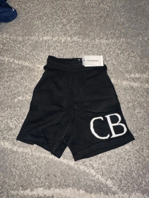 Svarta stickade shorts från Cole Buxton - Svarta stickade shorts från Cole Buxton med hög midja och bred resår. Stora vita CB-bokstäver på ena benet och två rejäla bakfickor. Perfekt för dig som vill ha en chill och trendig look.