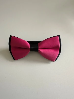 Helt NY satin-fluga i rosa och svart - Festlig dubbelknäppt fluga i rosa och svart satin med justerbart band för bästa passform. Klassisk tidlös modell som passar till alla festliga tillfällen. Helt ny oanvänd. 