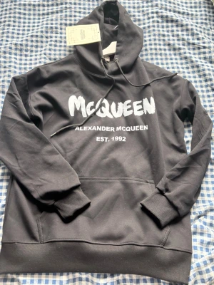 Svart hoodie från Alexander McQueen - Snygg svart hoodie från Alexander McQueen med stor vit logga och texten 'EST. 1992' på bröstet. Klassisk känguruficka framtill, ribbade muddar och justerbar huva med snören. Perfekt för dig som vill ha en streetwear vibe och gillar exklusiva märken.