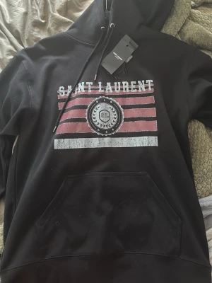 Svart hoodie från Saint Laurent - Snygg svart hoodie från Saint Laurent med tryck framtill i vitt och rött samt texten 'SAINT LAURENT'. Hoodien har huva med dragsko och en stor magficka. Perfekt för dig som vill ha en streetwear-look med exklusiv känsla. Nytt skick, kom privat för bättre bilder- pris:1899 kan diskuteras vid snabb affär