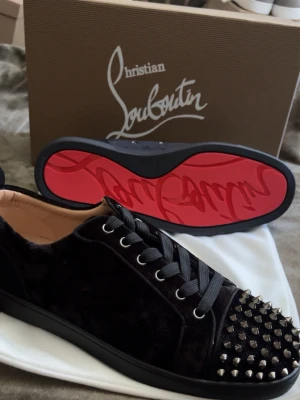 Christian Louboutin svarta sneakers - Svarta sneakers från Christian Louboutin med klassisk röd sula, svarta skosnören och silverfärgade nitar framtill. Ovandelen är i sammet och glitterdetaljer, vilket ger en lyxig och edgy vibe. Perfekta för dig som vill sticka ut med din stil.