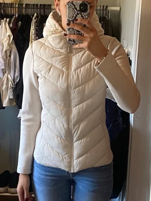 Vit quiltad pufferjacka med huva - Snygg vit pufferjacka med quiltad design och dragkedja framtill. Jackan har en stor, mysig huva och långärmade ärmar. Perfekt för kyliga dagar och ger en clean look till din outfit. Ena snöret vid luvan har gått av (se bild). 