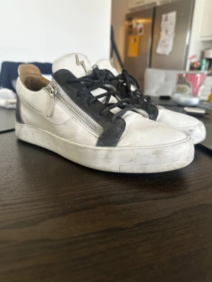 Svarta och vita sneakers från Giuseppe Zanotti - Snygga sneakers i vitt läder med svarta detaljer och svarta skosnören. Dragkedja på sidorna och ikonisk blixtlogga på plösen. Skorna har en låg siluett och en chunky vit sula. Perfekta för dig som gillar streetstyle och exklusiva märken.