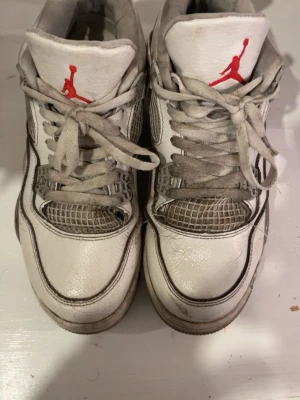Air Jordan 4 Oreo  - Säljer ett par vita Air Jordan 4 Oreo sneakers med röda Jumpman-loggor på plösen och grå detaljer. Skorna har meshpaneler, klassisk snörning och en robust sula. Tillverkade i läder och textil, perfekta för dig som gillar streetwear och ikoniska modeller.