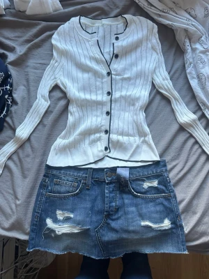 Mink jeanskjol + kofta - Säljer en outfit perfekt för sommaren och våren! Säljer en vit ribbad kofta med svarta knappar och svarta kantdetaljer. Koftan har lång ärm, rund halsringning och smal passform. Tröjan är väldigt stretchig och skulle gissa på att den passar från xs till storlek M! Toppen är använd 1 gång och säljs då den inte passar mig. Med outfiten följer en demin minikjol ifrån topshop! Kjolen har lappen kvar och är aldrig använd. Jeanskjolen är i storlek S/36!