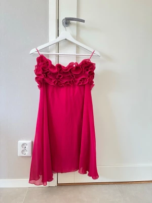 Rosa kortklänning med volang - Supersöt rosa kortklänning! Off-shoulder och stora volangdetaljer upptill. Klänningen har en luftig och flowy passform, perfekt för sommarens alla tillfällen. Materialet är  lätt och svalt.