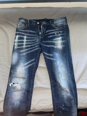 Blå jeans med slitningar och färgstänk - Säljer ett par blå DSQ2 jeans som är väldigt unik i modell och eftertraktad. Har endast använt 2 gånger själv men det tyvär inte är i min stil och vill sälja snabbt. Stl 32 men passar lite större samt mindre// kom pv för bättre bilder och frågor 