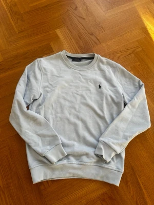 Ljusblå sweatshirt från Polo Ralph Lauren - Ljusblå sweatshirt från Polo Ralph Lauren med klassisk rund halsringning och broderad logga på bröstet. Tröjan har ribbade muddar vid ärmslut och nederkant. Perfekt för en clean och stilren look.
