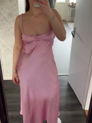 Rosa långklänning i satin - Säljs inte längre. Snygg rosa långklänning i satin med smala axelband och lätt rynk vid bysten. Klänningen har en mjuk glansig yta och en elegant, figurnära passform. Perfekt för dig som vill sticka ut med en feminin look. 