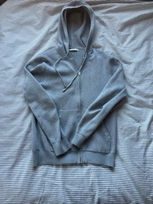 Ljusgrå zip-hoodie med fickor - Säljer en ljusgrå hoodie med dragkedja och huva. Tröjan har ribbad struktur, två framfickor och dragsko i huvan. Perfekt för en chill och avslappnad stil. Materialet känns mjukt och bekvämt mot huden.