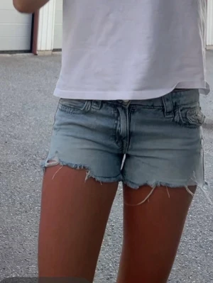 Ljusblå jeansshorts med fransig kant - Säljer ett par ljusblå jeansshorts med rå, fransig kant och slitna detaljer. Klassisk femficksmodell med knapp och dragkedja framtill. Perfekta till sommaren och ger en avslappnad vibe. Nypris || 250kr 