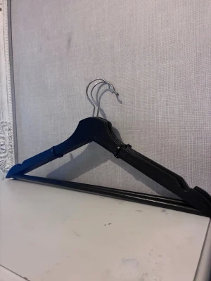 2st svarta galgar i trä - Två svarta galgar i trä med metallkrok, perfekta för att hänga upp kläder i garderoben. Galgarna har en klassisk form och är stabila med en stång längst ner för byxor eller kjolar.