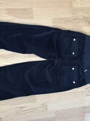 Svarta velourmjukisbyxor Juicy Couture - Säljer ett par svarta mjukisbyxor från Juicy Couture i velour med breda ben och dragsko i midjan. Byxorna har två stora fickor framtill och en diskret logga på ena fickan. Perfekta för chill och comfy vibes.