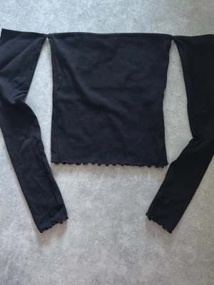 Svart off shoulder topp med volangkant - Trendig svart off shoulder topp med långa ärmar och volangkant vid ärmslut och nederkant. Perfekt att styla till jeans eller kjol för en cool look. Mjuk och stretchig känsla.