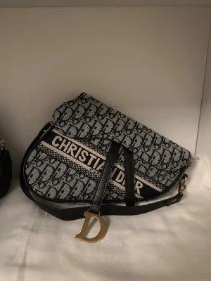 Christian Dior Saddle Bag  - Säljer en ikonisk Christian Dior Saddle Bag i svart och grått monogramtyg med tydlig logga och guldfärgad D-detalj på remmen. Väskan är perfekt inför sommaren och funkar nästan till alla outfits 💫🌐