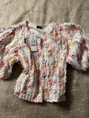 blommig topp - Superfin blus med volanger och puffärmar från Gina Tricot. Blusen är vit med blommigt mönster i rosa, blått och gult. Den har en strukturerad, rynkad yta och är perfekt för dig som gillar färg och statement-ärmar.