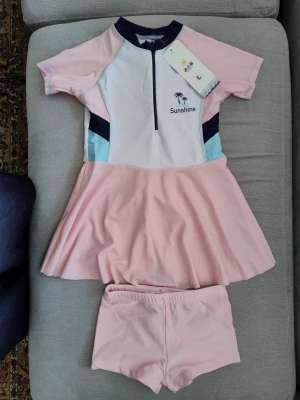 Rosa strandklänning med shorts Sunshine - Söt strandklänning i ljusrosa med matchande shorts från Sunshine. Klänningen har kort ärm, dragkedja framtill och detaljer i vitt, marinblått och ljusblått. Materialet är stretchigt och snabbtorkande, perfekt för stranden eller poolen.oanvänd  passar barn som är 4 åring