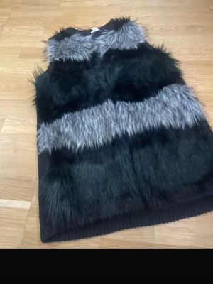Svart och grå pälsväst med fuskpäls - Snygg pälsväst i svart och grå fuskpäls med breda ränder och mjukt innerfoder. Västen är ärmlös och har en rak passform, perfekt för att lyfta din outfit med en trendig touch. Materialet är mjukt och fluffigt, vilket ger en cool och modern look.