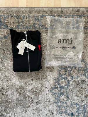 Ami Paris zip hoddie - Svart hoodie från Ami med dragkedja framtill och huva med snören. På bröstet finns ett rött broderat hjärta med A. Hoodien har två fickor och ribbade muddar. Materialet är mjukt och känns lyxigt, perfekt för en chill look.