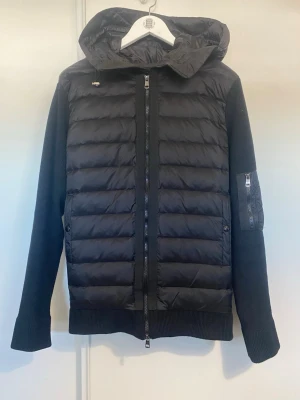 Svart pufferjacka med huva från Moncler - Snygg svart pufferjacka från Moncler. Jackan har stickade ärmar och rygg, dragkedja framtill och fickor med knappstängning. Detaljer som ficka med dragkedja på ärmen och Moncler-logga på axeln. Hör av er vid funderingar. 