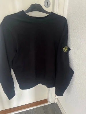 Svart sweatshirt från Stone Island - Cool svart sweatshirt från Stone Island med klassisk logotyp-patch på ärmen. Tröjan har rund hals och långa ärmar, perfekt för en avslappnad och trendig look. Materialet är mjukt och bekvämt, idealiskt för att matcha med jeans eller joggers.
