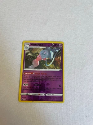 Pokémonkort Hatterene Psychic - Säljer ett Pokémonkort med Hatterene, Psychic-typ. Kortet har lila och gula kanter, med en färgglad illustration av Hatterene. Text och symboler i svart och vitt, samt detaljer om attack och förmåga. Perfekt för samlare eller Pokémonfans.