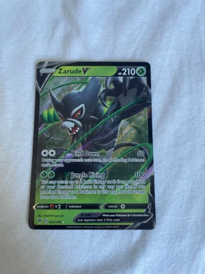 Pokémonkort Zarude V - Säljer ett Zarude V Pokémonkort med 210 HP från Sword & Shield-serien. Kortet har en färgstark design med Zarude i svart och grönt, och attacker som Bind Down och Jungle Rising. Perfekt för samlare eller Pokémon TCG-spelare.
