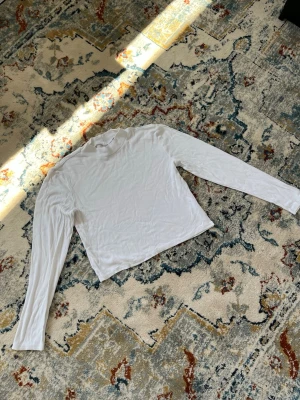 Vit ribbad långärmad topp - En stilren vit långärmad topp i ribbat material. Toppen har en högre krage och är figurnära i modellen, perfekt att styla med jeans eller kjol. Enkel och tidlös design som passar till många olika looks.