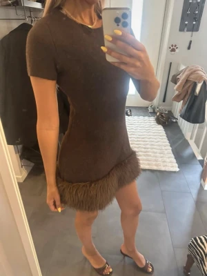 Brun kort klänning med fuskpäls från Zara - Snygg brun kortärmad klänning från Zara i stickat material med fuskpälsdetalj längst ner. Endast använd en gång. Klänningen har en fin passform och är perfekt för dig som vill sticka ut med en unik detalj. Supersnygg till klackar eller boots!