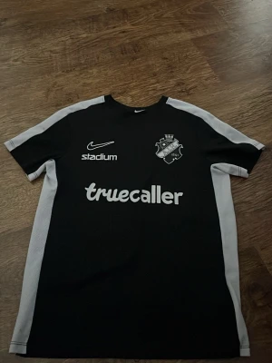 Svart AIK tränings t-shirt Nike - Svart tränings t-shirt från Nike med AIK:s klubbmärke på bröstet. T-shirten har vita detaljer längs sidorna och axlarna samt tryck från Truecaller, Stadium, Fabege och SVEA. Perfekt för fotbollsträning eller sportiga aktiviteter.