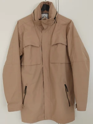 Vindjacka från McKinley i nyskick! - Stilren beige vindjacka med hög krage och dragkedja. Jackan har två stora bröstfickor med lock och två snedställda fickor med dragkedja. Jackan har även en ficka för att gömma luvan "syns på bild" Lång modell med minimalistisk design, perfekt för blåsiga dagar. Priset kan diskuteras vid snabb affär.