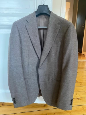 Brun ullkostymkavaj med klassiska slag - Snygg brun/grå kavaj med två knappar och klassiska slag. Kavajen har två framfickor och en bröstficka, samt knappar vid ärmsluten. Märket är Volt - These Glory Days Nypris: 2499kr Nyskick - helt oanvänd.  Material: ull 100%