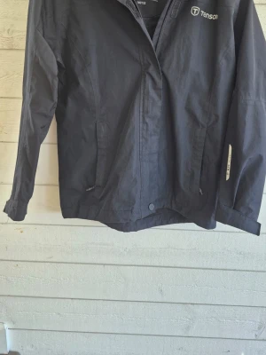 Svart skaljacka från Tenson - Snygg svart skaljacka från Tenson med huva och dragkedja framtill. Jackan har två fickor med dragkedja, diskret logga på bröstet och reflexdetalj på ärmen. Perfekt för blåsiga och regniga dagar och enkel att matcha med olika outfits.