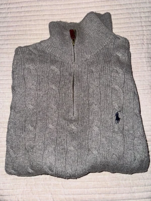 Grå kabelstickad tröja Polo Ralph Lauren - Kabelstickad grå tröja från Polo Ralph Lauren med halv dragkedja och hög krage. Tröjan har det klassiska broderade logotypen på bröstet och ribbade muddar. Perfekt för lager-på-lager och chill stil.