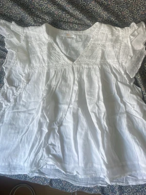 Vit blus med volangärmar - Säljer en luftig vit blus med v-ringning och volangdetaljer på axlarna. Blusen har korta ärmar och är gjord i ett tunt, lätt material som känns skönt mot huden. Perfekt för dig som gillar en romantisk och avslappnad stil.