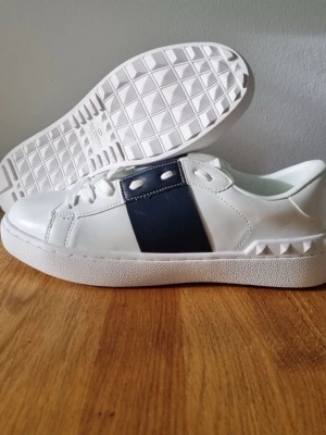 Valentino Open sneakers med blå detalj - Snygga vita Valentino Open sneakers med en bred marinblå panel på sidan och coola nitar längs hälen. Skorna har snörning och platt sula, tillverkade i skinn för en lyxig känsla. Perfekta för dig som vill ha en stilren och trendig look.