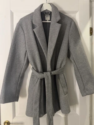 Ljusgrå kappa från ONLY med skärp - Stilren ljusgrå kappa från ONLY med klassisk krage och knytskärp i midjan. Kappan har raka ärmar och två sidofickor. Perfekt för dig som vill ha en snygg look! 