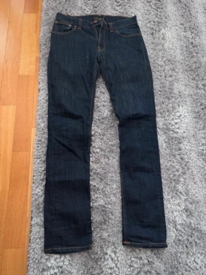 Mörkblå jeans från Nudie Jeans - Säljer ett par mörkblå jeans från Nudie Jeans med klassisk femficksmodell och kontrastsömmar. Jeansen har rak passform och är tillverkade i kraftig denim. Perfekta för dig som gillar stilrena och tidlösa jeans. W31 L34