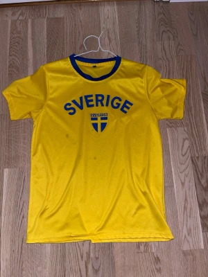 Gul Sverige t-shirt med blå detaljer - Gul t-shirt med blå text 'Sverige' och svensk flagga på bröstet. Rund halsringning med blå kant. Perfekt för att visa ditt stöd för Sverige. Materialet känns som polyester och har en sportig look.