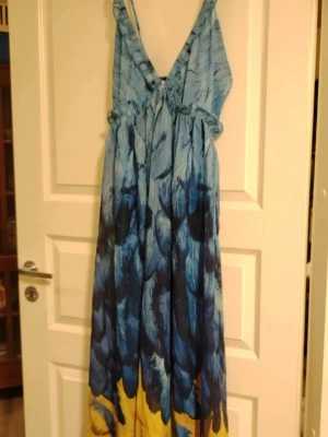 Blå maxiklänning med fjädermönster🩵  - Lång maxiklänning med djup v-ringning och smala axelband. Klänningen har ett blått fjäderinspirerat mönster med inslag av gult nertill. Materialet känns lätt och luftigt, perfekt för varma dagar☀️ Snygg volangdetalj längs ringningen❤️ S-M. 