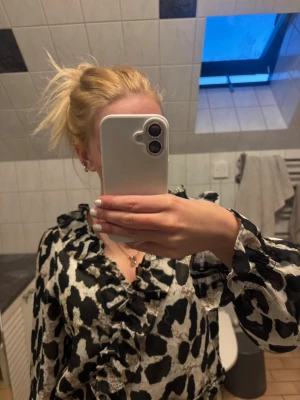 Leopardmönstrad volangblus - Säljer en snygg blus med svart och beige leopardmönster. Blusen har volangdetaljer längs ringningen och framtill, samt långa, lätt puffiga ärmar med resår vid ärmslut och midja. Materialet är tunt och lite transparent, vilket ger en luftig känsla.
