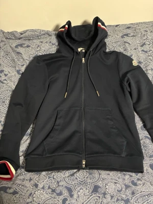 Mörkblå Moncler hoodie med dragkedja - Snygg mörkblå hoodie från Moncler med hel dragkedja, fickor fram och huva med rödvita detaljer. Ärmslut och huva har Monclers signaturrand i rött, vitt och blått. Klassisk logga på ärmen. Perfekt för en stilren och sportig look.