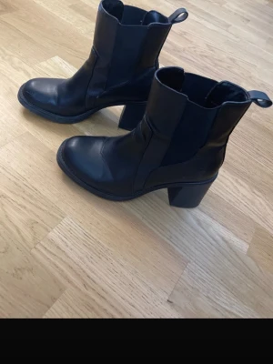 Svarta stövletter med klack - Säljer ett par svarta stövletter i skinnimitation med bred klack och rundad tå. De har dragflikar upptill och resår på sidorna för enkel påtagning. Snygg och stilren modell som passar till många outfits.