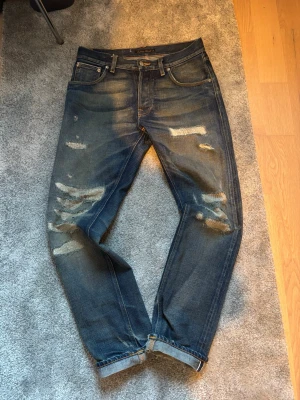Slitna Nudie Jeans - Säljer ett par as schyssta slevedge denmin Nudie jeans. Dem är extremt unika med slitningar och lagade detaljer på benen, dem har även fått en speciell tvätt under tidens gång. Dem har en rak passform och sitter snyggt. Vill du ha ett par unika Nudies är dem för dig. 