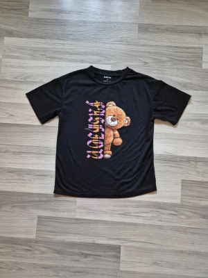 Svart t-shirt med nalletryck från SHEIN - Cool svart t-shirt från SHEIN med ett stort tryck av en brun nalle och färgglad text på framsidan. T-shirten har klassisk passform och korta ärmar, perfekt för att ge din outfit en lekfull touch.