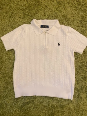 Vit kabelstickad piké från Polo Ralph Lauren - Vit kabelstickad pikétröja från Polo Ralph Lauren med kort ärm, krage och halv dragkedja framtill. Klassisk logga broderad på bröstet. Perfekt för en clean och stilren look.