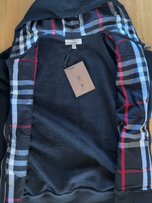 Svart hoodie från Burberry med rutigt foder - Svart hoodie från Burberry med klassiskt rutigt foder i rött, vitt och blått. Hoodien har dragkedja och huva, samt mjukt innerfoder. Perfekt för dig som vill ha en stilren och exklusiv look med ikoniskt Burberry-mönster på insidan.